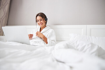 Young pretty lady in white plush bathrobe using modern device in bedroom