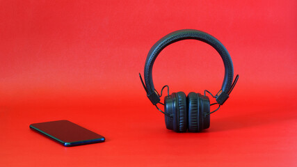 Wireless headphones with ear pads next to a smartphone on a red background. The concept of listening to music, radio and audio blogs using wireless technology. Free space for an inscription