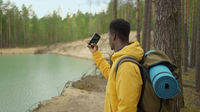 A black man with a backpack uses a mobile phone while hiking through the forest in the mountains across the lake. Take a photo and blog