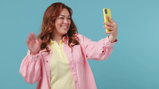 Young pretty redhead chubby overweight woman 20s years old in pink jacket yellow t-shirt doing selfie shot on mobile phone post photo on social network isolated on pastel blue color background studio