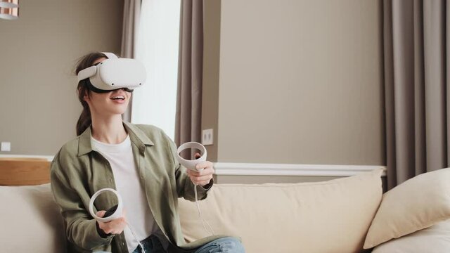 Happy young woman with virtual reality headset or 3d glasses playing games sitting on sofa at home