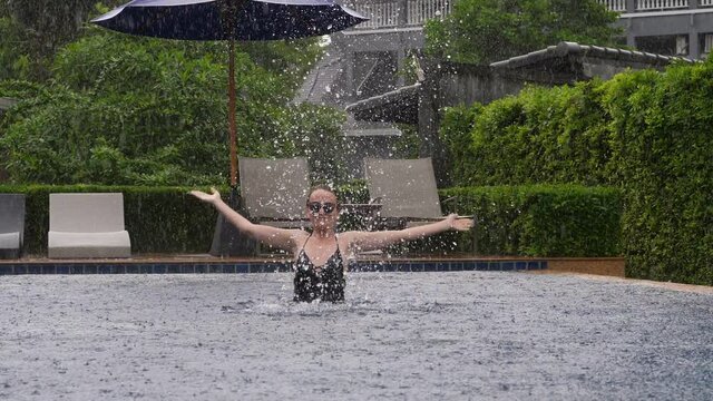 Young adult woman play in swimming pool, she stand in water and jump up, throw water up and towards camera by hands. Then move down and spring again with hands to sides, slow motion shot