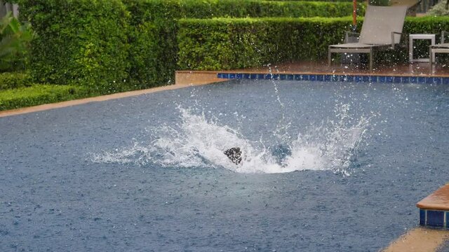 Adult woman clumsily jump in swimming pool, create big splash of water. Slow motion shot of tourist have fun at rainy day, fall plop in pool, heavy tropical rain pouring down