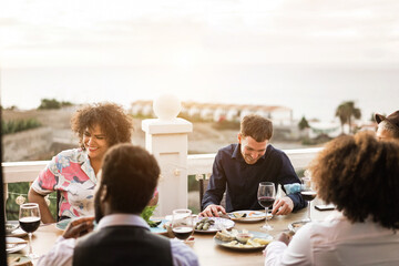 Happy multiracial friends having dinner and drinking wine outdoor at home - Focus on faces