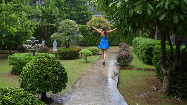 Woman enjoy warm tropical rain, raise up hands and stroll away at green garden, view from back. Typical daily shower at rainy season in Thailand, tourist lady have fun despite of weeping skies