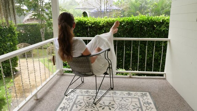 Nowhere to run at rainy day, woman sit in chair at villa porch and put legs on railings. She relax, listen rain noise of and look to tropical shower pouring down at green garden