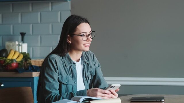 The smiling woman with glasses texting on the phone and looking forward while sitting at the table in the kitchen