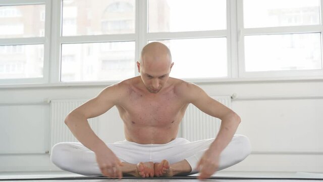Experienced yogi male performing Baddha Konasana - Butterfly Pose sitting on fitness mat, shirtless bald man concentrated on workout. Yoga, sport, wellbeing concept.