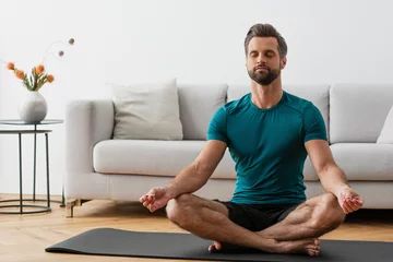 Fototapeten man with closed eyes meditating in lotus pose on yoga mat © LIGHTFIELD STUDIOS