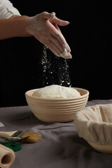 Cropped view of female hands flouring dough ball in ribbed rattan basket for bread proofing. Proofing basket with linen liner cloth, parchment and cooking brush are located on gray tablecloth. 