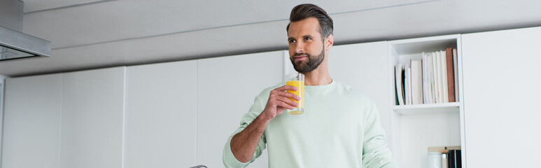 joyful man looking away while holding glass of orange juice in kitchen, banner
