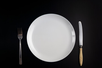 Fork, knife and white plate on black background in flat lay