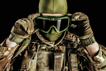 Photo of soldier in level 3 armored vest ammunition, tactical gloves putting on tactical goggles on black background.