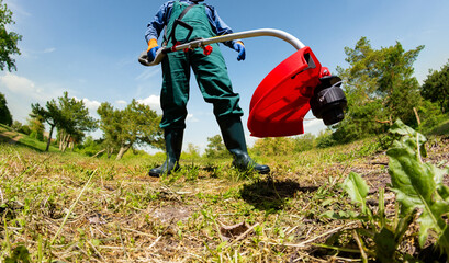 Photo of worker in green overalls and rubber boots using electric garden trimmer and cutting grass.