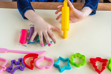 Girl toddler holds colorful play clay by one hand and yellow toy rolling pin by other hand. Play dough mold set for enhancing fine motor skills. Kid education creativity using toy play doh