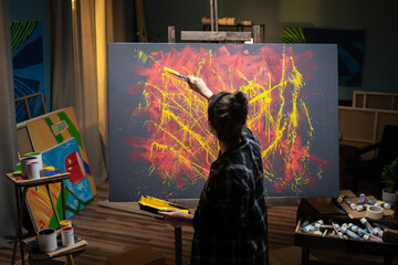 The artist spends time in the studio painting an abstract painting in tones of yellow and red. The female holds a paint brush in front of her and splashes yellow paint across the canvas.