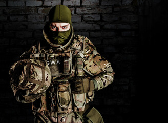 Photo of fully equipped swat soldier in heavy level 3 amor ammunition and rifle standing on dark bricks background.