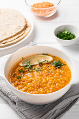 Masoor dal. Indian spicy lentil soup in white plate, roti (chapati), sliced green coriander and dry raw lentil are on white wooden table.