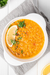 Masoor dal. Indian spicy red lentil soup in white plate,  half a lemon, sliced green coriander, gray napkin are on white wooden table.