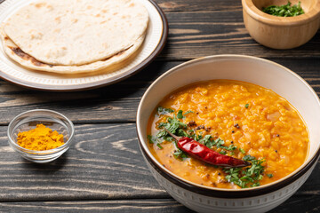 Masoor dal. Indian spicy red lentil soup, roti (chapati), sliced green coriander, and dry raw lentil are on brown wooden table.