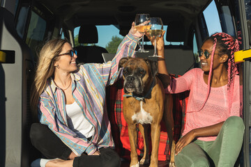 Two girls of different ethnicities toasting with wine inside a van with a boxer dog in the countryside.