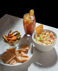 Tuna salad with elbow macaroni mayonnaise strips of carrots jalapeño or jalapeno chili and tortilla chips and an ice tea on the saide over a white table, li by the sunlight