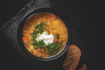 Soup with pickled cucumbers and pearl barley in black bowl on black wooden table background