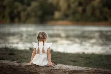 a cute little girl in the summer sits on log on the river bank in a white dress and looks into the distance. nature, vacation, walk in the park. selective focus. safety of children by the water, pond