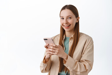 Smiling young attractive girl using mobile phone, chatting on smartphone and looking at camera pleased, standing over white background