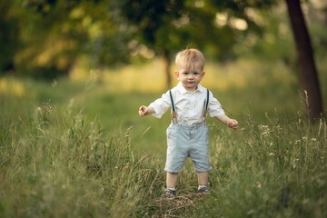 cute little blond toddler boy in a white shirt and suspenders is walking in the park, smiling....