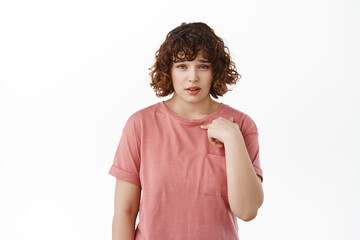 Are you talking about me. Surprised and doubtful woman pointing at herself with disbelief, being chosen or accused, showing hesitation, standing against white background