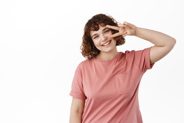 Positive cute girl smiling happy, showing peace kawaii v-sign and gazing at camera, standing lovely against white background in t-shirt