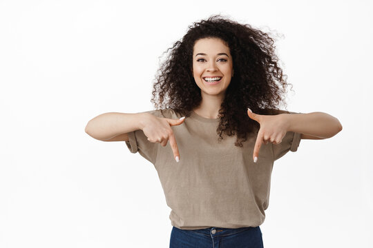 Portrait Of Brunette Girl With Curly Hair And White Teeth, Smiling While Pointing Fingers Down, Inviting To Event, Click This Link, Showing Logo Below, Standing Against White Background