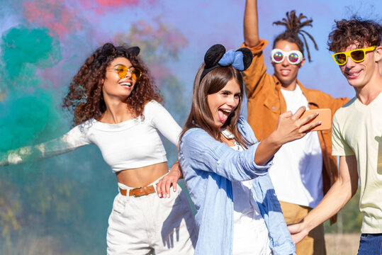 Beautiful young woman hold smartphone, doing selfie with happy friends, having fun in the park with multicolored smoke bombs smoke fog. Young students celebrating spring break together. Holi festival.