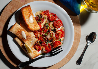 A cherry tomato snack on a white plate with toasted bread with butter and a fork on the side over a wooden cutting board and a glass cup of olive oil, a spoon and a blue napkin. Olive oil dressing