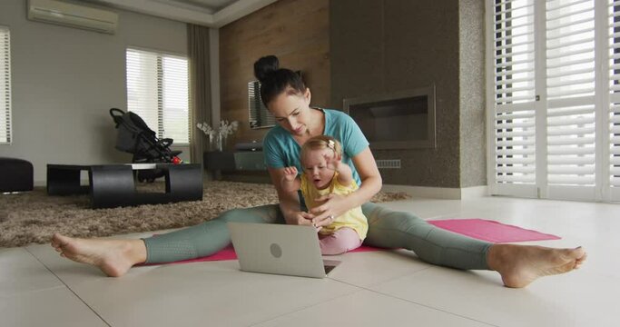 Caucasian mother holding her baby practicing yoga on yoga mat in front of laptop at home