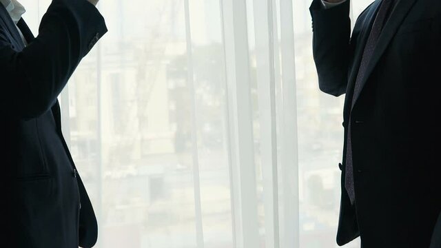 Silhouette of two men in business suits clinking cognac glasses. Toasting against window. Detail of businessmen hands raising toast after deal.
