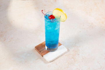 A view of a blue colored alcoholic beverage, in a highball glass, garnished with a cherry and lemon...