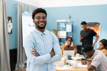 Portrait of a smiling dark-skinned man wearing a shirt with his arms crossed over his chest, the guy has dark hair and stubble, a business meeting of a group of employees is going on in the background