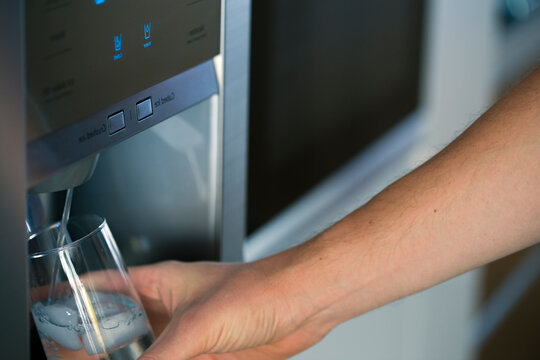 Male hand is getting cold water with ice cube from dispenser of home fridge. Concept of convenience.