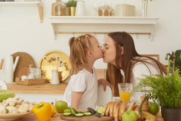 Cheerful mother and little daughter are preparing salad together in the kitchen and having fun. The girl kisses her mother at home