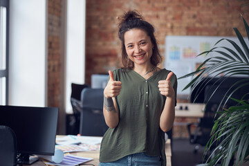 Portrait of cheerful woman, interior designer or architect smiling at camera, showing thums up while standing in modern green office on a daytime