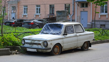 Old rusty white passenger car with flat wheels in the courtyard of a residential building, Malookhtinsky Prospekt, Saint Petersburg, Russia, May 2021