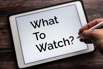 Tablet displaying “What To Watch” on white background. Man hand holding wireless stylus pen in image. Holiday watch movie at home or cinema with digital gadget or device concept
