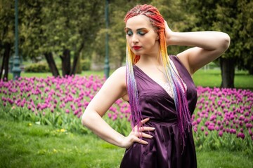 A beautiful girl with expressive makeup and African braids in a bright purple dress walks in a blooming park near large flower beds with tulips
