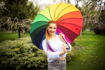 A cute girl with multi-colored braids and bright makeup in a bluish shirt posing with a rainbow umbrella against the background of a blooming spring park