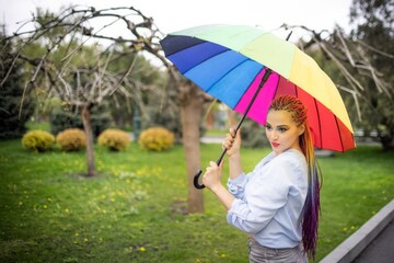 A cute girl with multi-colored braids and bright makeup in a bluish shirt posing with a rainbow umbrella against the background of a blooming spring park