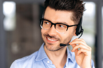 Close up portrait of successful joyful young caucasian operator or call center manager wearing glasses, in formal attire, using headset for video conference or phone call, looking away, smiling