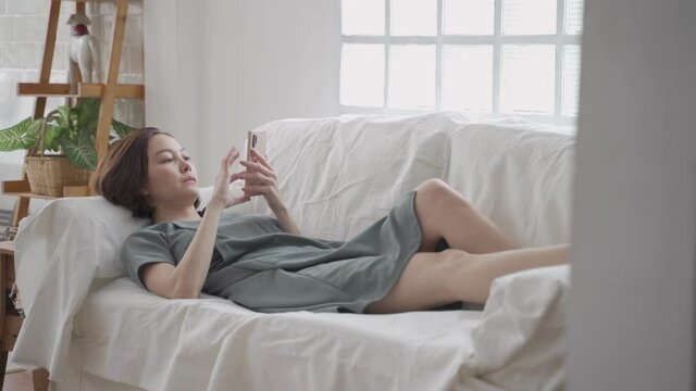 candid shot, asian woman laying down on sofa couch relaxing using mobile smart phone in living room at minimal style home. female holding mobile phone concentrate on reading looking on text.