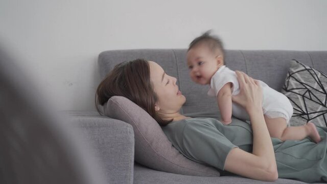 asian woman mother holding happy baby infant playing and kissing on sofa in living room at minimal style home in day time. candid authentic shot life at home. mom play with baby girl by love and care.